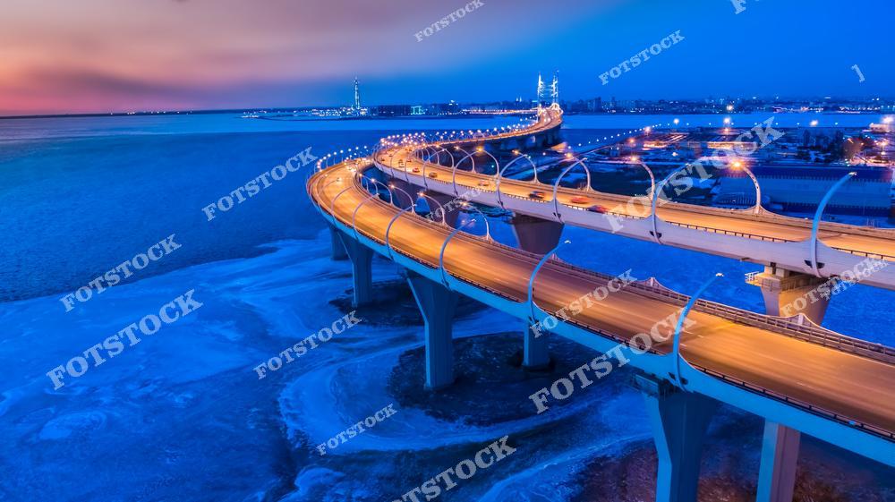 Панорамная фотография промышленного района Санкт-Петербурга зимой, снятая с высоты птичьего полета. На снимке видны современные автомобильные магистрали, транспортные развязки, промышленные предприятия и окраины города, уходящие к побережью Финского залива. Зимний городской пейзаж подчеркивает масштаб инфраструктуры, развитие транспортной сети и индустриальную архитектуру Северной столицы России. Вид с дрона позволяет оценить геометрию дорог, движение транспорта и особенности промышленной зоны Санкт-Петербурга. Фотография отлично подходит для публикаций о городской инфраструктуре, логистике, промышленности, урбанистике, фотостоков и рекламных проектов.